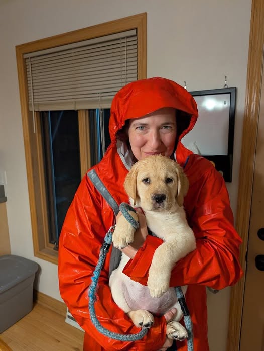 Person in an orange raincoat holding a yellow Labrador puppy indoors, smiling at the camera.